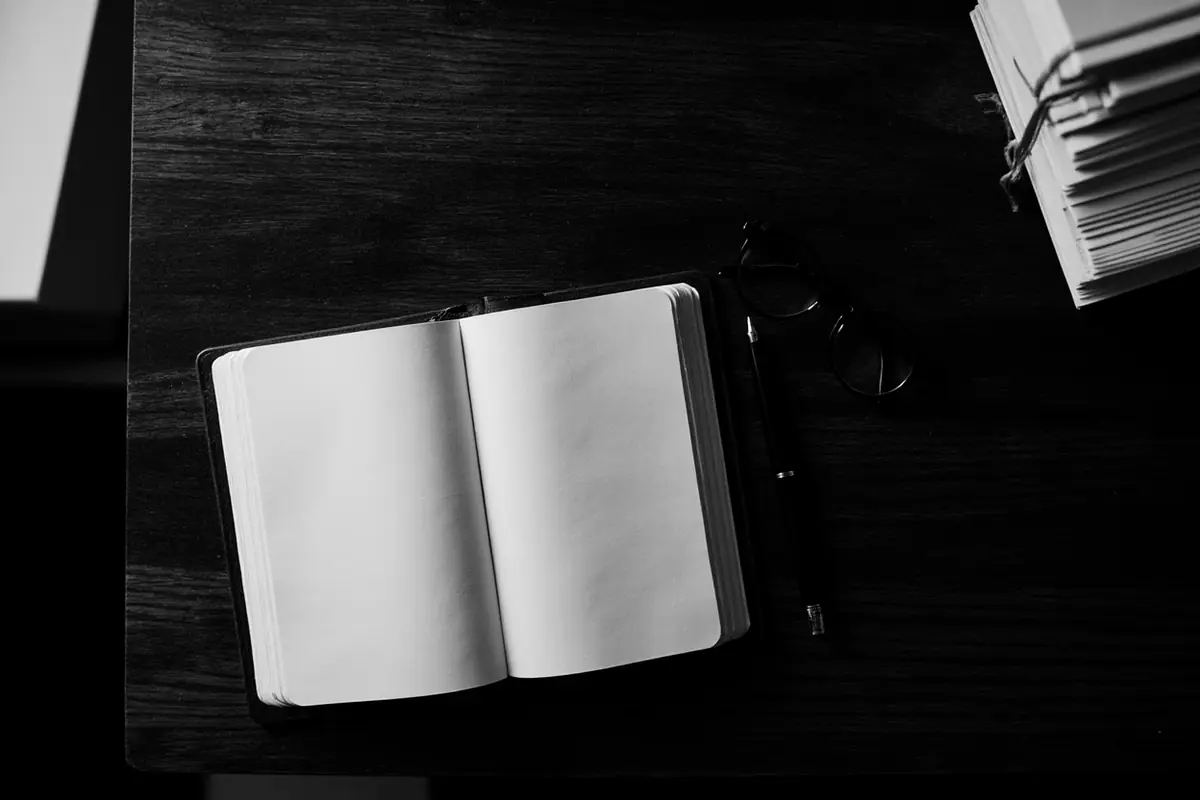 Koyu ahşap çalışma masasında açık defter, dolma kalem, gözlük ve bağlanmış dosya yığını